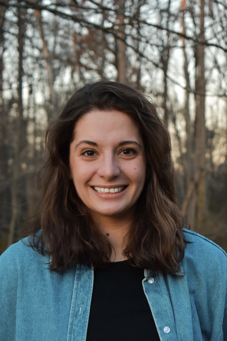 A color photo of Milena, a smiling woman with shoulder-length brown hair, wearing a black T-shirt and a denim jacket. She is standing in front of a blurred background of autmnal trees.