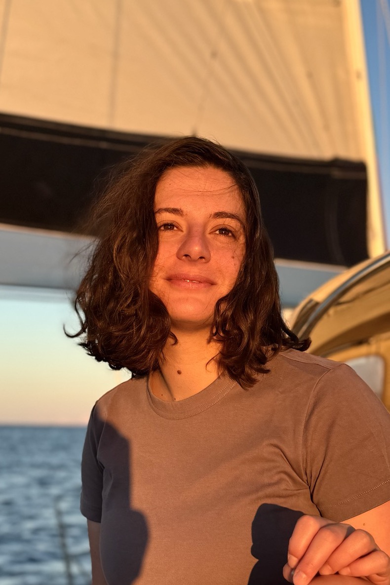A color photo of Milena, a woman with shoulder-length wavy brown hair and a soft smile, wearing a brown T-shirt. She is sitting on a sailboat in warm golden-hour light, with a white sail behind her and the sea in the background.